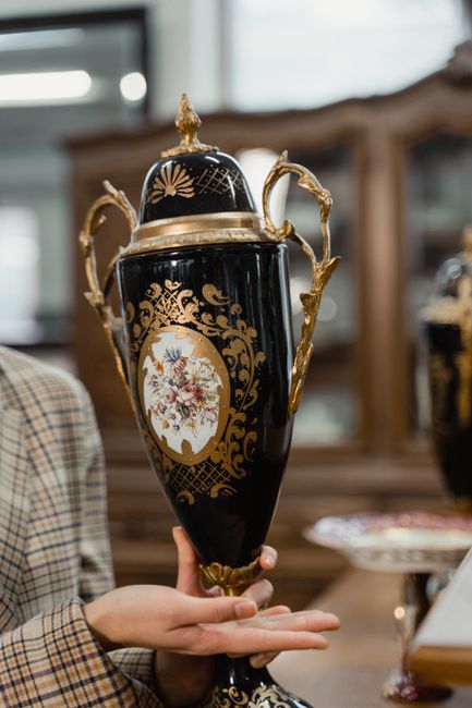 A person with a checkered blazer is holding an ornate black and gold porcelain vase with floral decorations and gold handles during a home relocation or moving process. The vase is small and delicate,